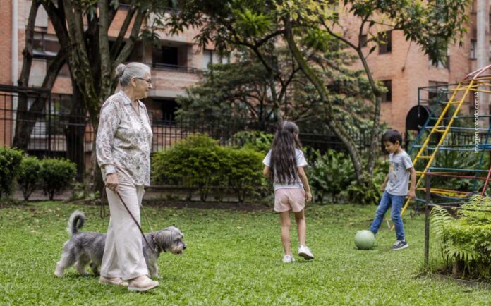 Persona mayor pasea con su perro mientras niños juegan en una zona verde residencial, imagen que representa el cuidado y la convivencia en Patio Bonito.
