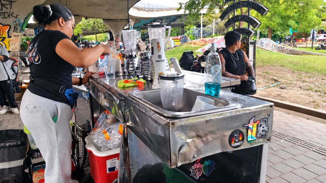 Mujer prepara bebidas en un puesto de jugos bajo el puente de la 4 Sur, con licuadoras sobre el mostrador y zonas verdes al fondo.