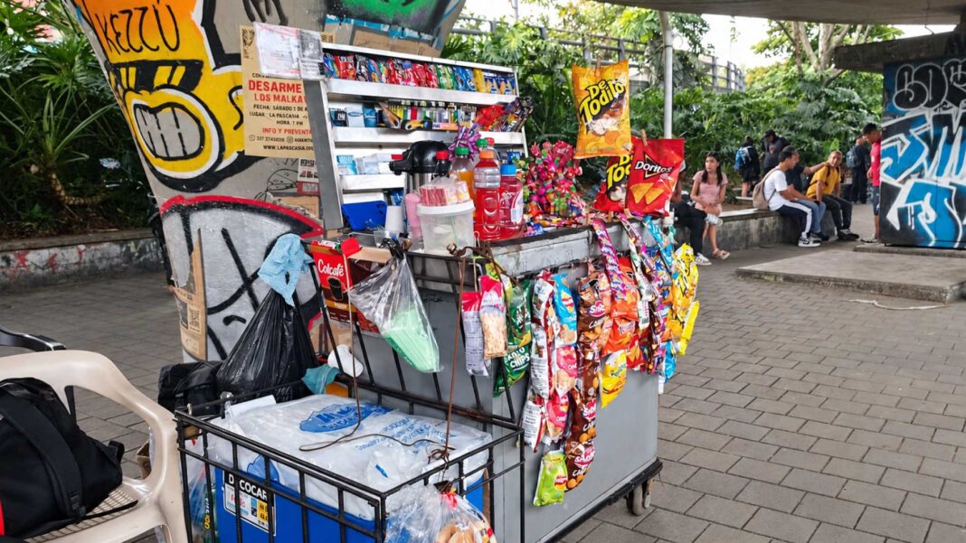 Carrito de dulces, bebidas y pasabocas bajo el puente de la 4 Sur, junto a columnas con grafitis y personas sentadas al fondo.